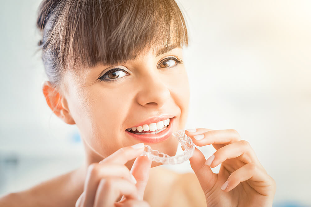 Woman smiling at camera and holding her clear Invisalign retainer