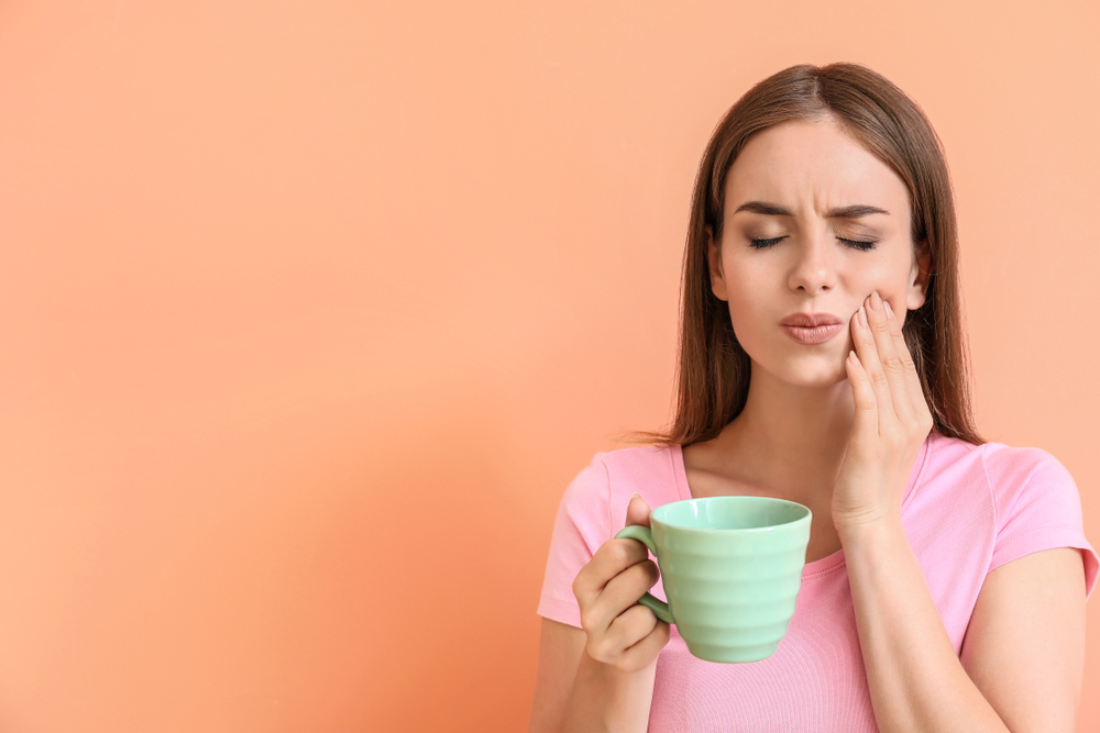 Young woman holding coffee cup and touching cheek in pain