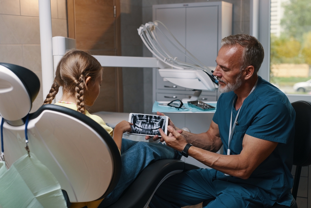 Pediatric dentist showing dental X-ray on a tablet to young girl during consultation in a modern dental clinic